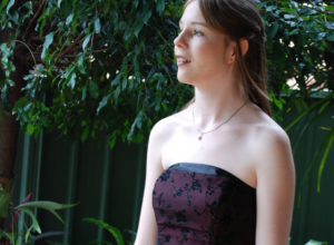 A 17-year-old Jayne is ready for her year 12 formal; she's wearing a red formal dress with black lace standing in front of a green garden