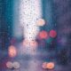A blurred city street at dusk is behind an in-focus pane of glass with rain droplets